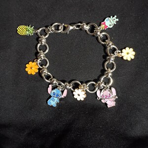 May include: Silver-toned charm bracelet featuring colorful enamel charms. Charms include a pineapple, flowers, and cartoon characters in blue and pink. The bracelet is on a black background.