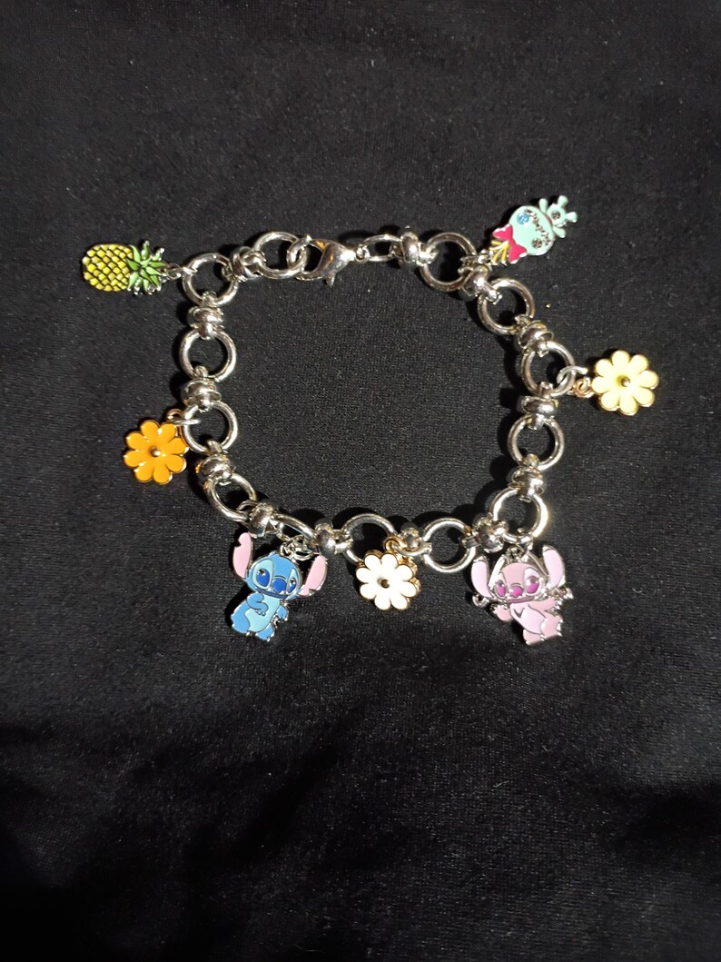 May include: Silver-toned charm bracelet featuring colorful enamel charms. Charms include a pineapple, flowers, and cartoon characters in blue and pink. The bracelet is on a black background.