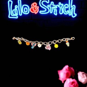 May include: A gold-colored charm bracelet featuring colorful enamel charms of cartoon characters and a pineapple. The bracelet is displayed against a black background with a neon sign that reads "Lilo & Stitch".