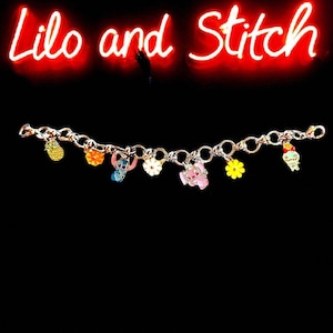 May include: A gold-colored charm bracelet featuring colorful enamel charms of characters and objects from the animated film "Lilo and Stitch." The charms include a pineapple, flowers, and the characters Stitch and Angel. The words "Lilo and Stitch" are written in red neon.