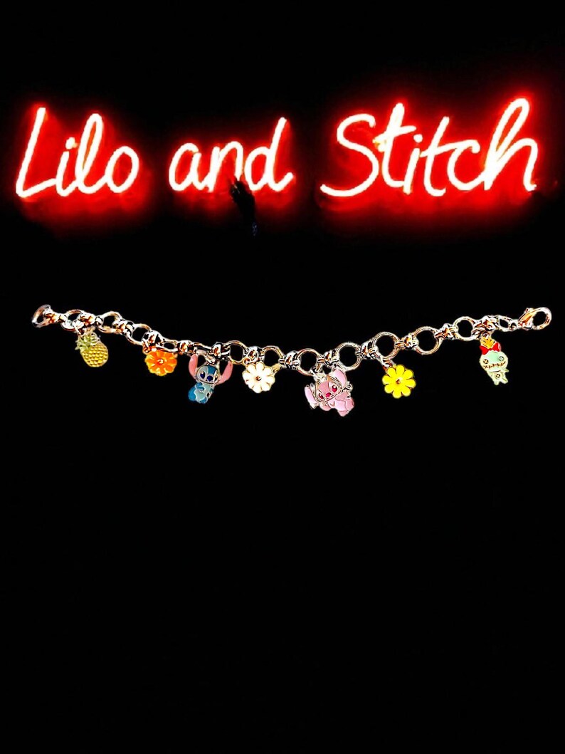 May include: A gold-colored charm bracelet featuring colorful enamel charms of characters and objects from the animated film "Lilo and Stitch." The charms include a pineapple, flowers, and the characters Stitch and Angel. The words "Lilo and Stitch" are written in red neon.