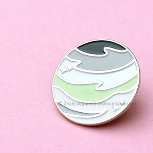 May include: A round, silver-coloured pin with a green, white, grey, and black design. The design resembles a planet with stripes. The pin has a white background with small white stars.