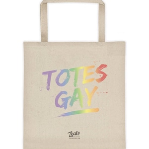 Puede incluir: Una bolsa de tela beige con dos asas a juego. La parte frontal presenta el texto TOTES GAY en una fuente de estilo pincelada con los colores del arcoíris. Debajo del texto, un pequeño logotipo negro dice Zealo Apparel.