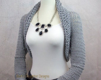 May include: A gray, knitted bolero sweater with long sleeves and a wide collar. The sweater is displayed on a mannequin, paired with a white tank top, dark jeans, and a silver and black statement necklace.