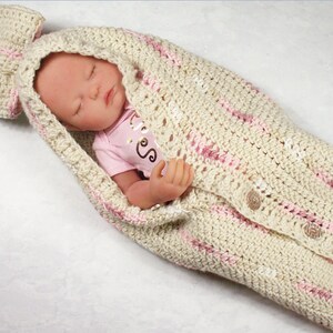 May include: A crocheted baby swaddle with a pink bow on the hood. The swaddle is cream colored with pink stripes and has buttons down the front.