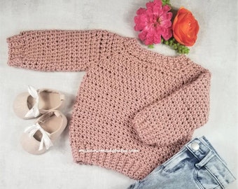 Puede incluir: Un suéter de punto rosa con mangas largas, colocado sobre una superficie blanca con un par de zapatos blancos y unos vaqueros azules. El suéter tiene un diseño sencillo y está hecho con un hilo suave.
