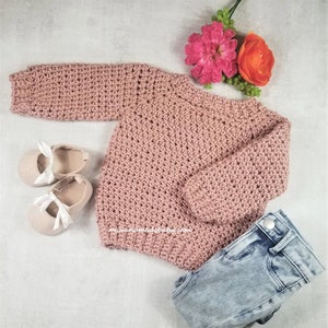 Op de afbeelding: Een roze gebreide trui met lange mouwen, gelegd op een wit oppervlak met een paar witte schoenen en een blauwe spijkerbroek. De trui heeft een eenvoudig ontwerp en is gemaakt van zacht garen.