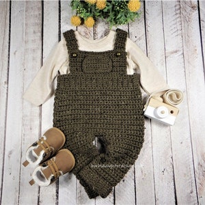 May include: A handmade olive green crochet baby overall with a matching long-sleeved shirt and brown baby shoes. The overall has suspender straps and decorative buttons. A small wooden toy camera is also present.