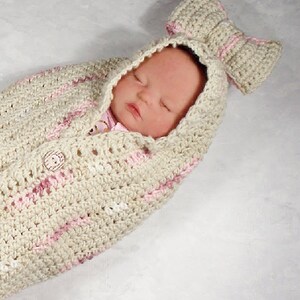 May include: A crocheted baby swaddle with a pink and white striped pattern and a matching crocheted bow. The swaddle has a button closure and a hood.
