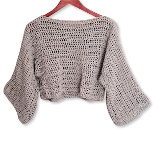 May include: A light taupe crochet sweater with wide, bell sleeves. The open weave design is visible throughout the cropped top. The sweater is displayed on a wooden hanger against a white background. The garment has a boat neck.