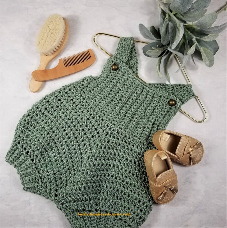 May include: A sage green crocheted baby romper with two gold-colored baby shoes. The romper has a button closure and is hanging on a gold hanger.