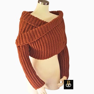 May include: A terracotta-coloured, off-the-shoulder crochet shrug. The garment has long sleeves and a wide, folded collar. The crochet stitch creates a textured appearance. The shrug is displayed on a mannequin against a white background.