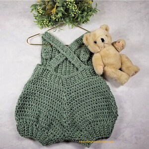 May include: A sage green crocheted baby romper with a criss-cross strap design. The romper is hanging on a gold hanger and a teddy bear is lying next to it.