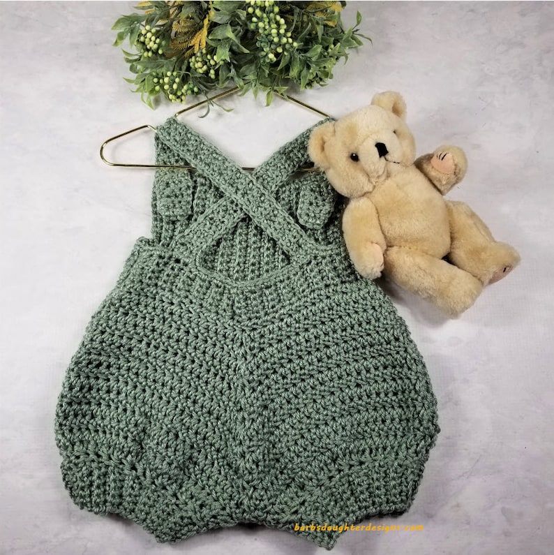 May include: A sage green crocheted baby romper with a criss-cross strap design. The romper is hanging on a gold hanger and a teddy bear is lying next to it.