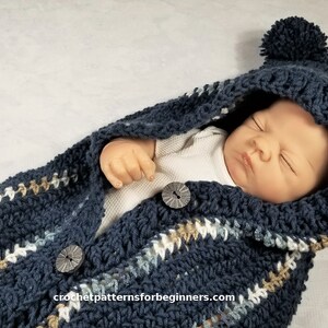 May include: A blue crocheted baby sleeping bag with a hood and pom-poms. The sleeping bag has a striped pattern in white, beige, and blue. The sleeping bag has buttons down the front.
