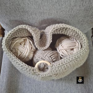 May include: A handmade, light gray crocheted yarn basket filled with several balls of yarn in shades of cream and gray. The basket has a wooden ring detail and a handle. The basket is sitting on a gray upholstered chair.