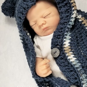 May include: A blue crocheted baby hooded wrap with white and tan stripes and pom poms on the hood. The wrap is buttoned with a silver button. The wrap is worn by a sleeping baby.