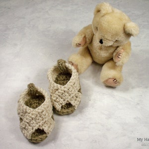 May include: Two pairs of crocheted baby sandals in beige and brown. The sandals are on a white surface with a teddy bear sitting behind them.