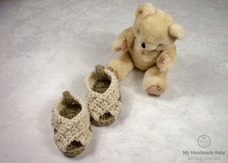 May include: Two pairs of crocheted baby sandals in beige and brown. The sandals are on a white surface with a teddy bear sitting behind them.