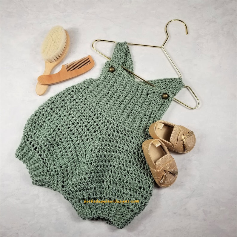 May include: A sage green crocheted baby romper with two gold buttons. The romper is hanging on a gold hanger. There are two pairs of tan leather baby shoes with tassels.