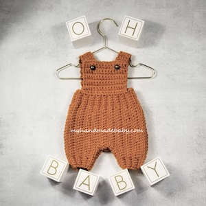 May include: A handmade, rust-coloured crochet baby romper with two small buttons on the straps, hanging on a silver hanger. White blocks spell out "OH BABY" around the romper. The website "myhandmadebaby.com" is printed on the romper.