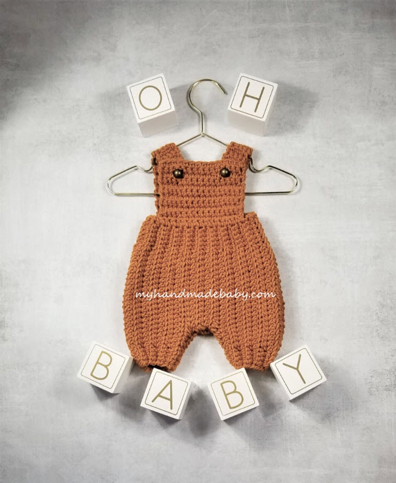 May include: A handmade, rust-coloured crochet baby romper with two small buttons on the straps, hanging on a silver hanger. White blocks spell out "OH BABY" around the romper. The website "myhandmadebaby.com" is printed on the romper.