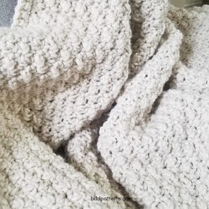 Puede incluir: Primer plano de una manta de ganchillo de color crema. La manta está hecha con un punto texturizado y tiene un aspecto suave y acogedor.