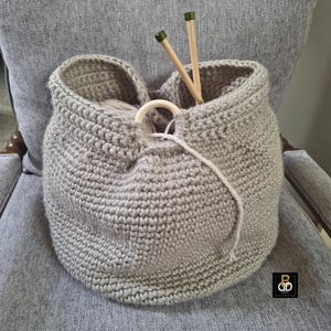 May include: A gray, crocheted yarn basket with a wooden ring detail and two wooden knitting needles. The basket is filled with yarn and sits on a gray upholstered chair. The basket is handmade.