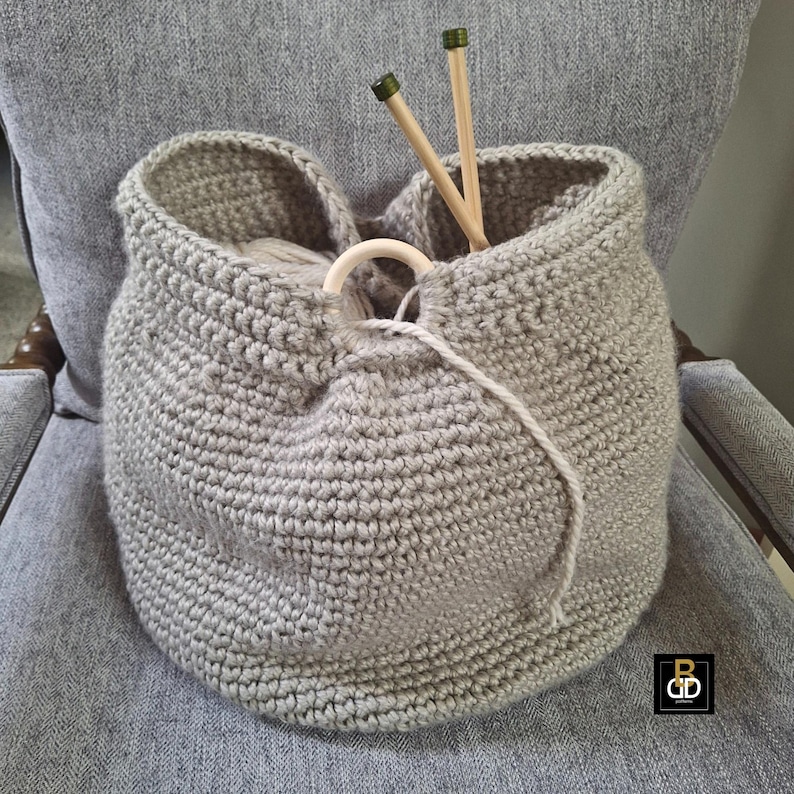 May include: A gray, crocheted yarn basket with a wooden ring detail and two wooden knitting needles. The basket is filled with yarn and sits on a gray upholstered chair. The basket is handmade.