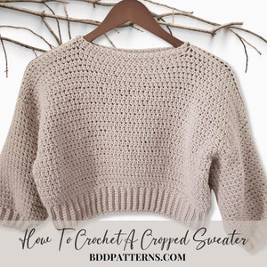 May include: A beige crocheted cropped sweater with long sleeves hanging on a wooden hanger. The sweater is made with a simple stitch pattern and has a relaxed fit. The text "How To Crochet A Cropped Sweater BDDPATTERNS.COM" is at the bottom of the image.