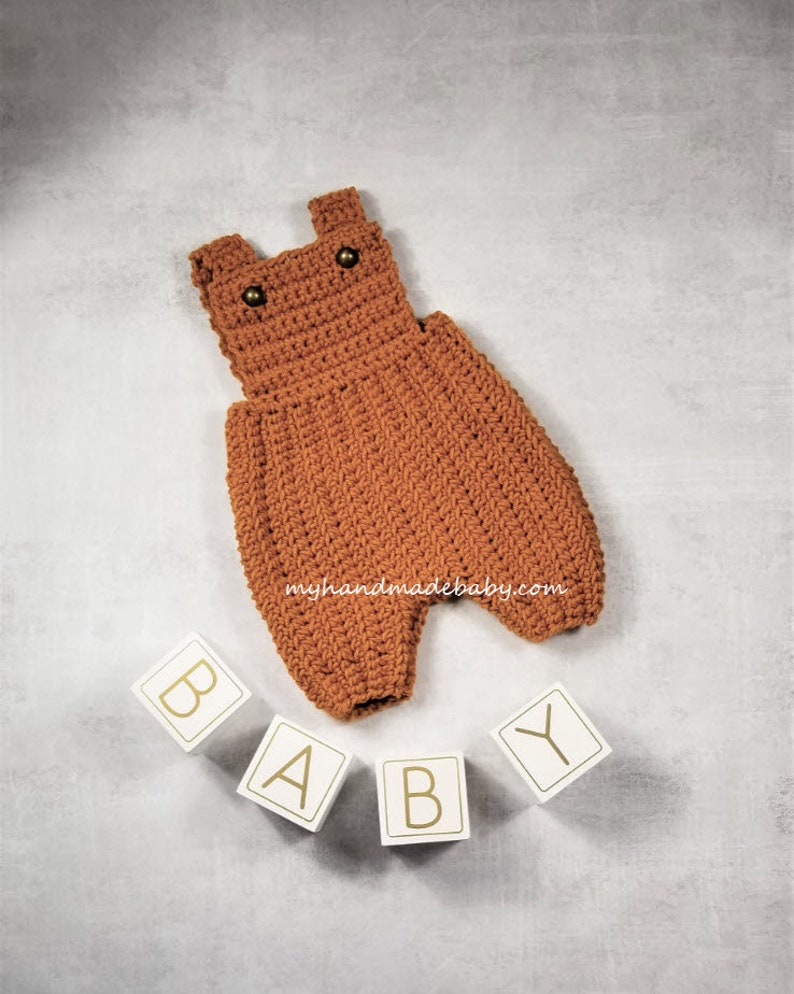 May include: A handmade, crochet baby romper in a warm, earthy brown. The romper features a classic overall design with button closures and a textured stitch pattern. The word "BABY" is spelled out with wooden blocks. The website address "myhandmadebaby.com" is visible.