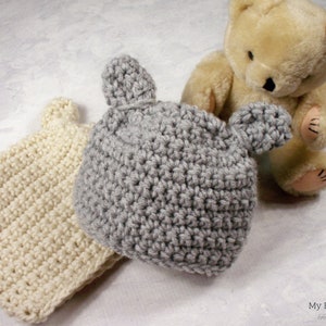 Puede incluir: Dos gorros de bebé de ganchillo con orejas de oso. Un gorro es de color crema y el otro es gris.