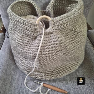 May include: A light gray, handmade crochet basket with a wooden ring handle. The basket is filled with yarn and a crochet hook is visible. The basket is sitting on a gray chair.