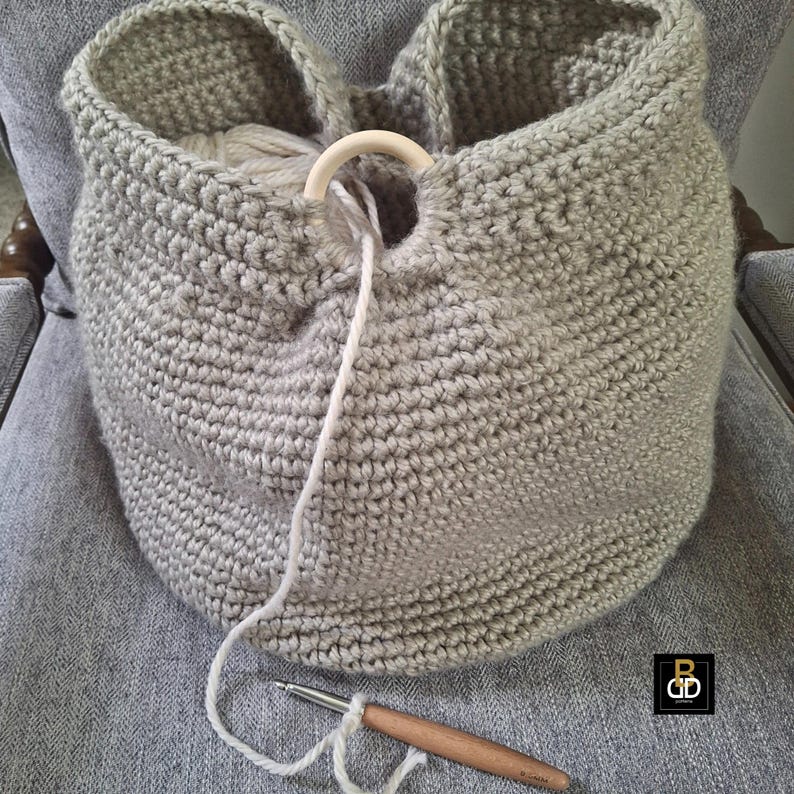 May include: A light gray, handmade crochet basket with a wooden ring handle. The basket is filled with yarn and a crochet hook is visible. The basket is sitting on a gray chair.