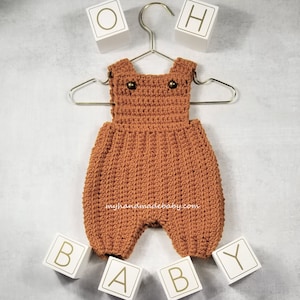May include: A handmade, russet-coloured crochet baby romper with suspender straps and button details, hanging on a gold hanger. The text "myhandmadebaby.com" is visible. White blocks spell "OH BABY".