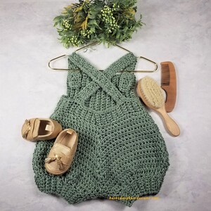 May include: A sage green crocheted baby romper with a criss-cross back design. The romper is hanging on a gold hanger and is accompanied by a pair of gold tassel loafers, a wooden hairbrush, and a wooden comb.