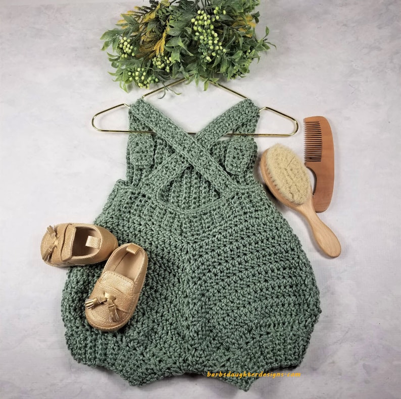 May include: A sage green crocheted baby romper with a criss-cross back design. The romper is hanging on a gold hanger and is accompanied by a pair of gold tassel loafers, a wooden hairbrush, and a wooden comb.
