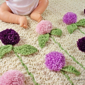 Op de afbeelding: Een gehaakt babydekentje met een witte achtergrond en een patroon van groene bladeren en kleurrijke pomponbloemen. De bloemen zijn in tinten roze, paars en lavendel.