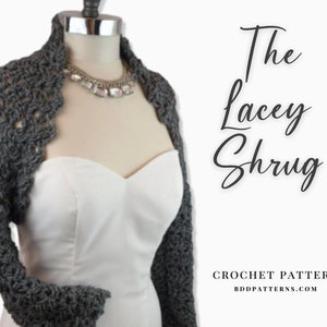 Può includere: Un bolero all'uncinetto grigio con bordo smerlato, esposto su un manichino. Il bolero è abbinato a un abito bianco e una collana d'argento. Il testo "The Lacey Shrug" e "CROCHET PATTERN" sono visibili.