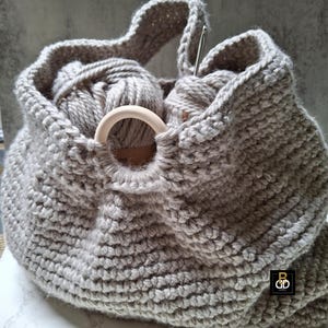 May include: A gray, hand-knitted yarn bag with a wooden ring detail. The bag is filled with gray yarn balls and a crochet hook. The bag has a handle for carrying and is designed for storing knitting or crochet supplies.