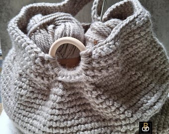 May include: A gray, hand-knitted yarn bag with a wooden ring detail. The bag is filled with gray yarn balls and a crochet hook. The bag has a handle for carrying and is designed for storing knitting or crochet supplies.