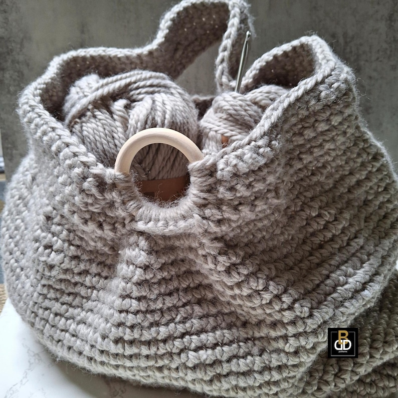 May include: A gray, hand-knitted yarn bag with a wooden ring detail. The bag is filled with gray yarn balls and a crochet hook. The bag has a handle for carrying and is designed for storing knitting or crochet supplies.