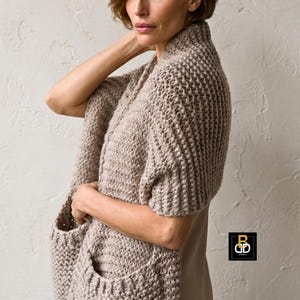 Può includere: Uno scialle beige a maglia grossa con due tasche frontali. Lo scialle è drappeggiato sulle spalle e sulle braccia. La maglia ha un aspetto strutturato e un colore neutro, adatto a vari outfit. Lo scialle è un accessorio comodo ed elegante.
