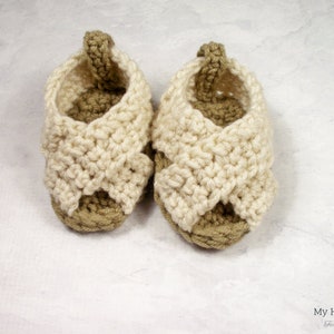 May include: A pair of crocheted baby sandals in beige and white. The sandals have a criss-cross strap design and are made with a soft yarn.