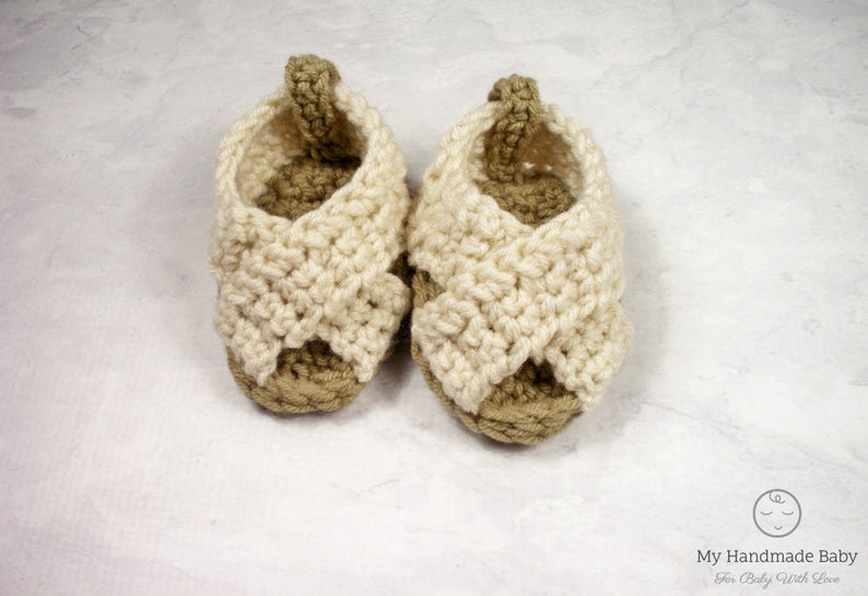 May include: A pair of crocheted baby sandals in beige and white. The sandals have a criss-cross strap design and are made with a soft yarn.