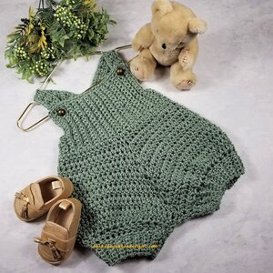 May include: A sage green crocheted baby romper with brown buttons. The romper is hanging on a gold hanger and is next to a brown teddy bear and two pairs of brown leather baby shoes.