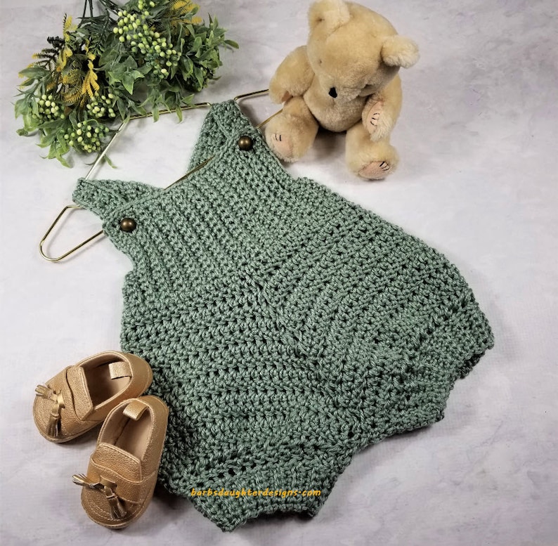 May include: A sage green crocheted baby romper with brown buttons. The romper is hanging on a gold hanger and is next to a brown teddy bear and two pairs of brown leather baby shoes.