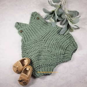 May include: A sage green crocheted baby romper with two gold buttons on the straps. The romper is lying on a white surface with two gold tassel loafers next to it.