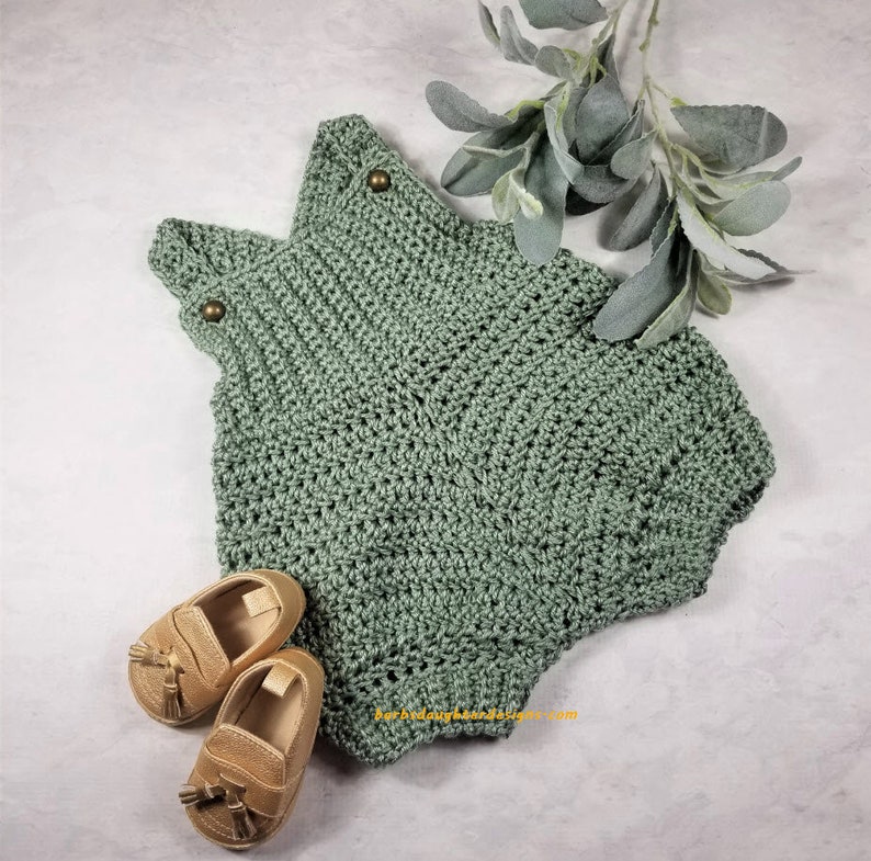 May include: A sage green crocheted baby romper with two gold buttons on the straps. The romper is laying on a white surface with two gold tassel loafers next to it.