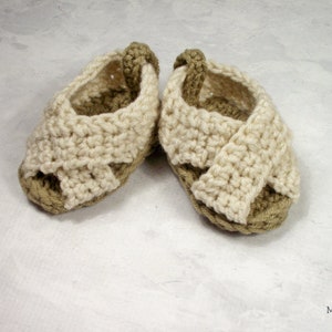 May include: A pair of crocheted baby sandals in beige and brown. The sandals have a criss-cross strap design and are open-toed.